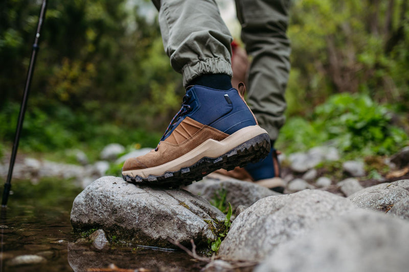 A close-up of a person hiking outdoors, stepping onto a rock in shallow water. The person is wearing olive green pants and a tan and navy blue hiking boot with a black sole.
