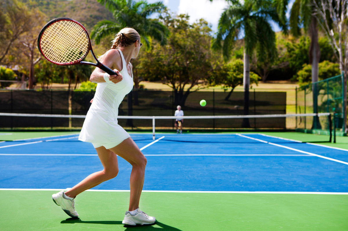 A woman in a white tennis dress swings a racket on a blue and green tennis court.