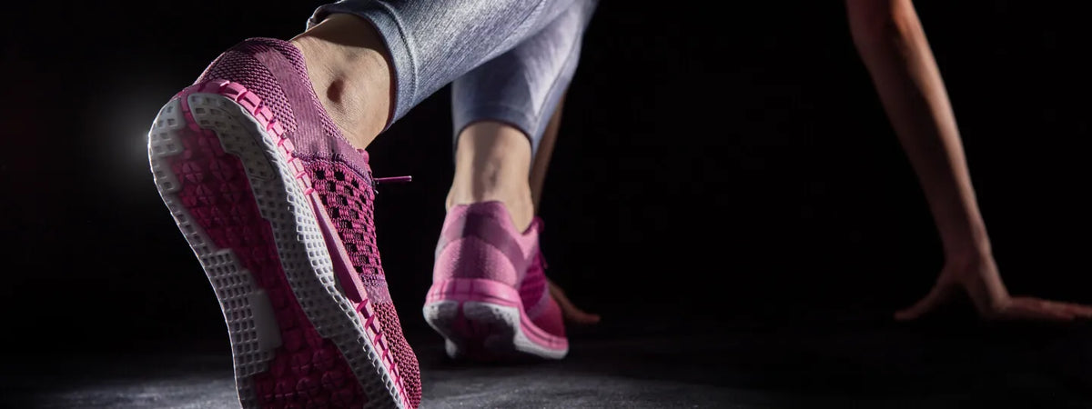 Low-angle close-up of a runner wearing pink mesh athletic shoes in a starting block position against a dark background.