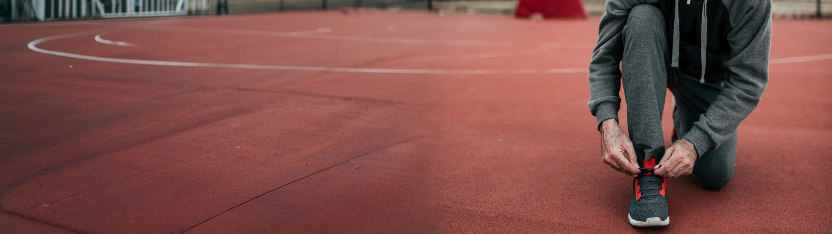 A person kneeling on a red running track, tying the laces of a black, gray, and red athletic shoe.