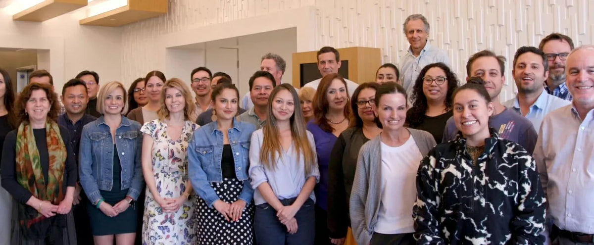 A large group of people posing together in an indoor office or meeting space with a textured white wall background.
